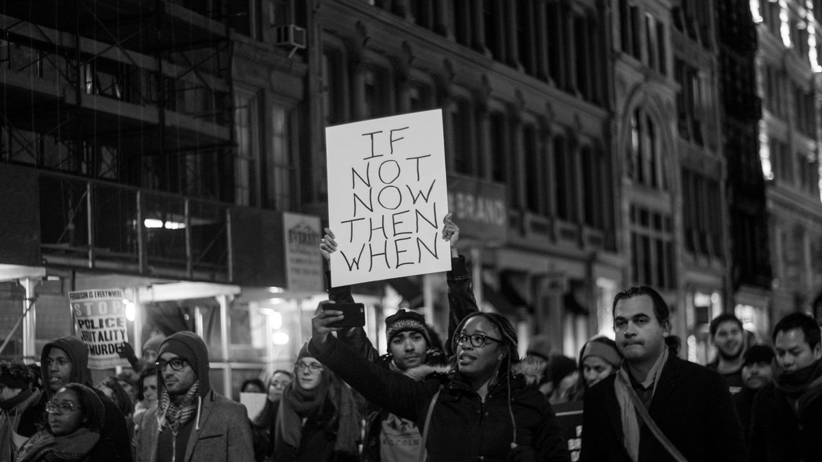 A marcher at a night protest holds a sign reading: If not now then when